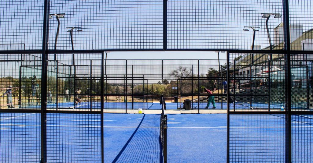 A wide-angle view of a bright blue outdoor padel court enclosed by black wire fencing and glass walls, with several people maintaining the court surface under a clear blue sky.