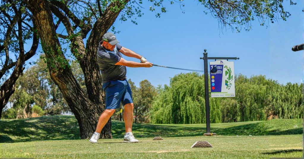 A male golfer in a grey shirt and blue shorts completing a powerful driver swing on a sunny grass tee box, with a course map sign and large trees in the background.